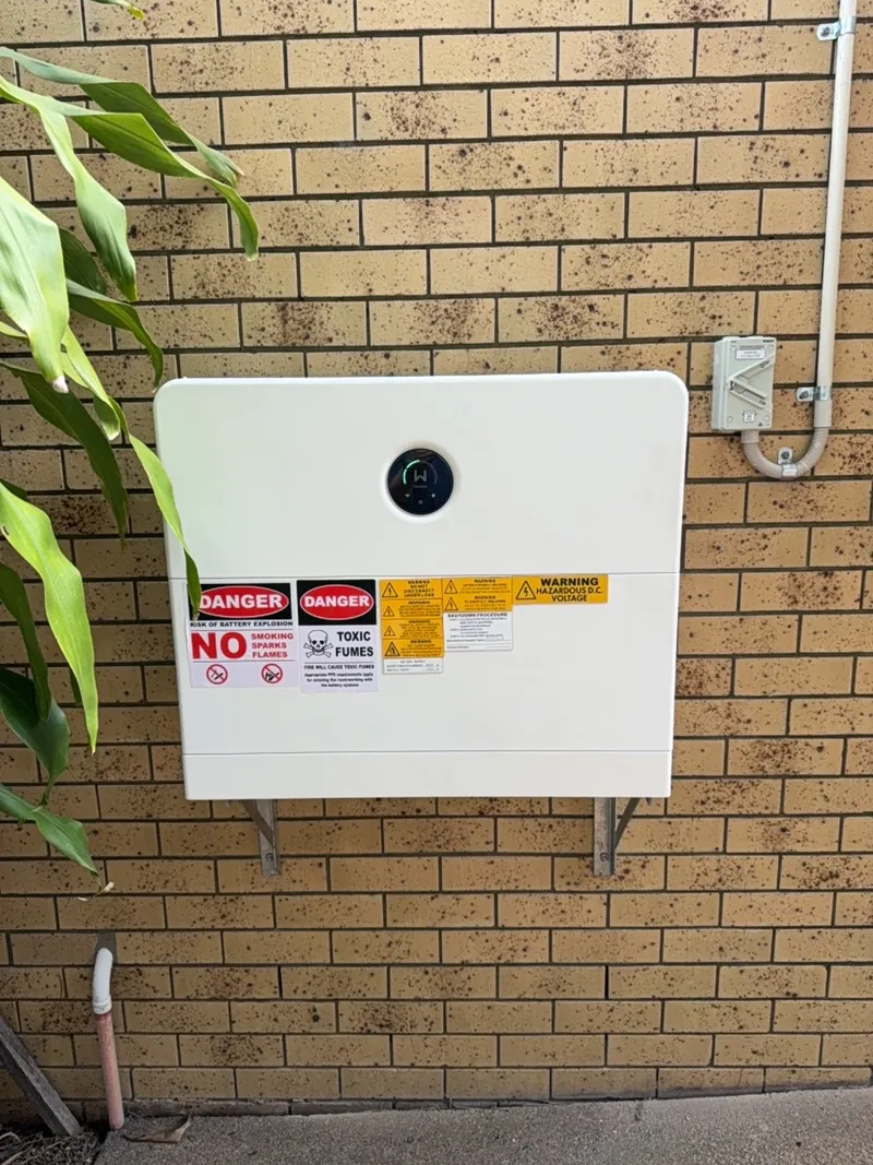 Weather-rated home battery mounted to an exterior brick wall with warning labels